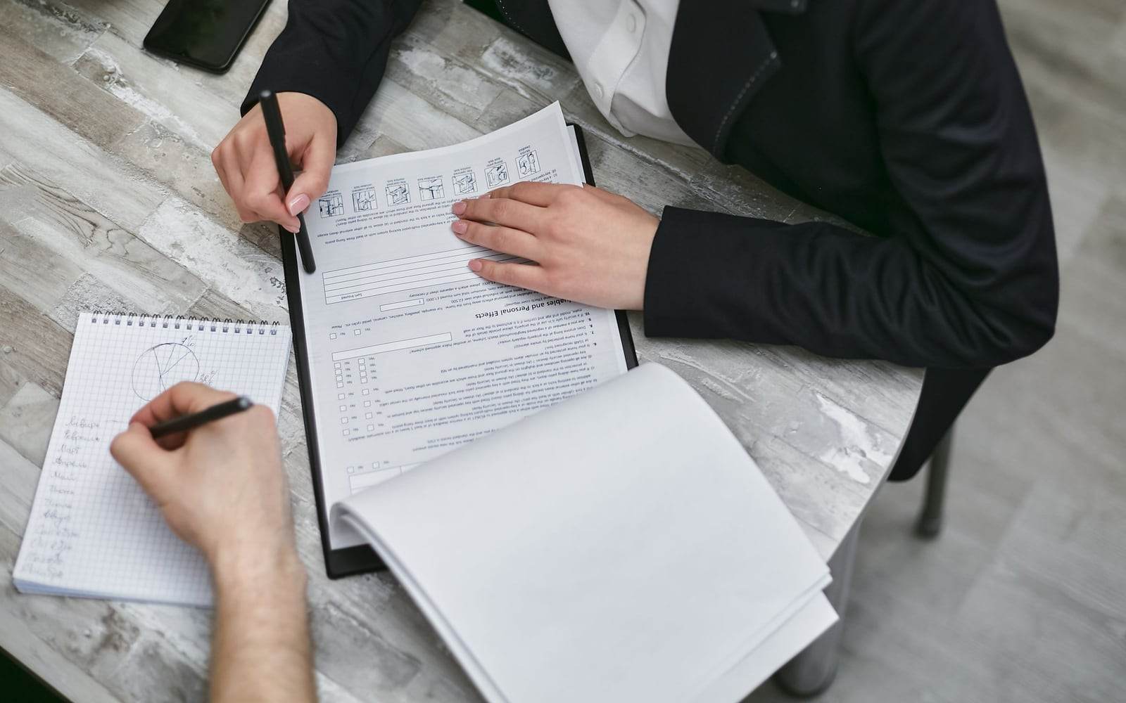 Two people review papers and take notes at a desk in a staged Pexels business photograph.